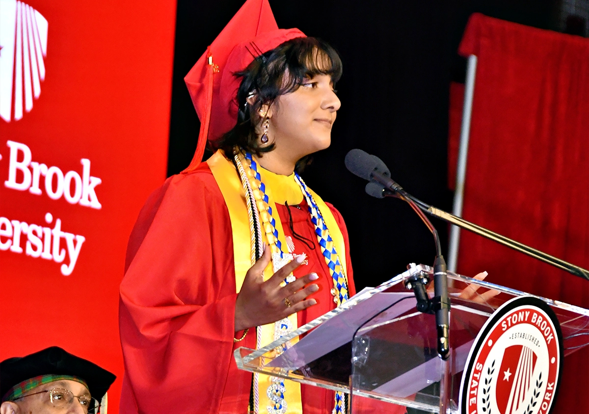 A Stony Brook University College of Business student, Karisma Persad, delivering a speech at a podium with bright red banners in the background