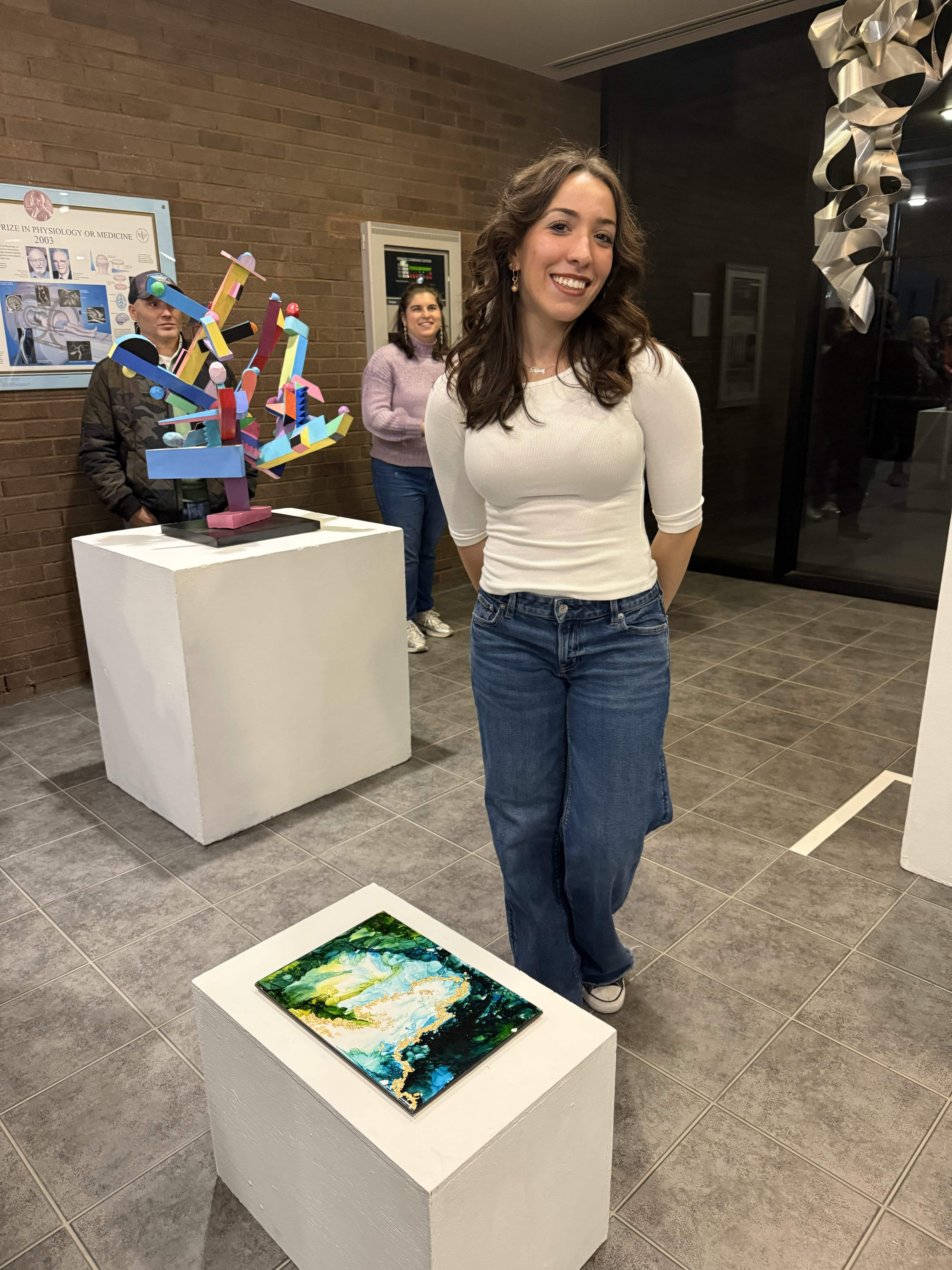 Art student, Eleni Tsevis wearing a white shirt and jeans stands next to her green and gold acrylic art sculpture