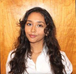 Portrait of a smiling individual with long curly hair, wearing a white shirt, against a wooden background.