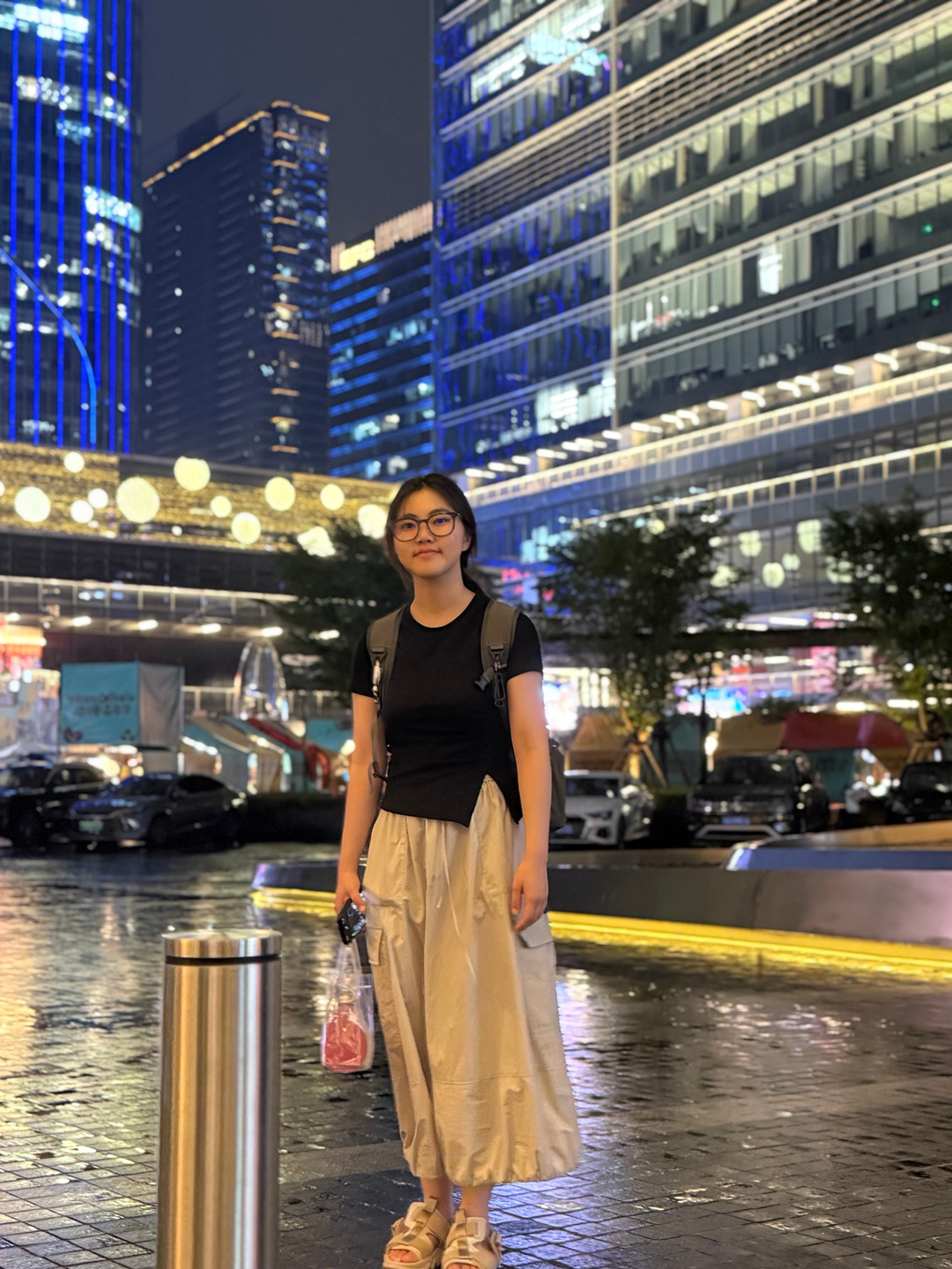 A person stands in front of brightly lit, modern skyscrapers at night, holding a water bottle and wearing glasses, a black top, and a long skirt.