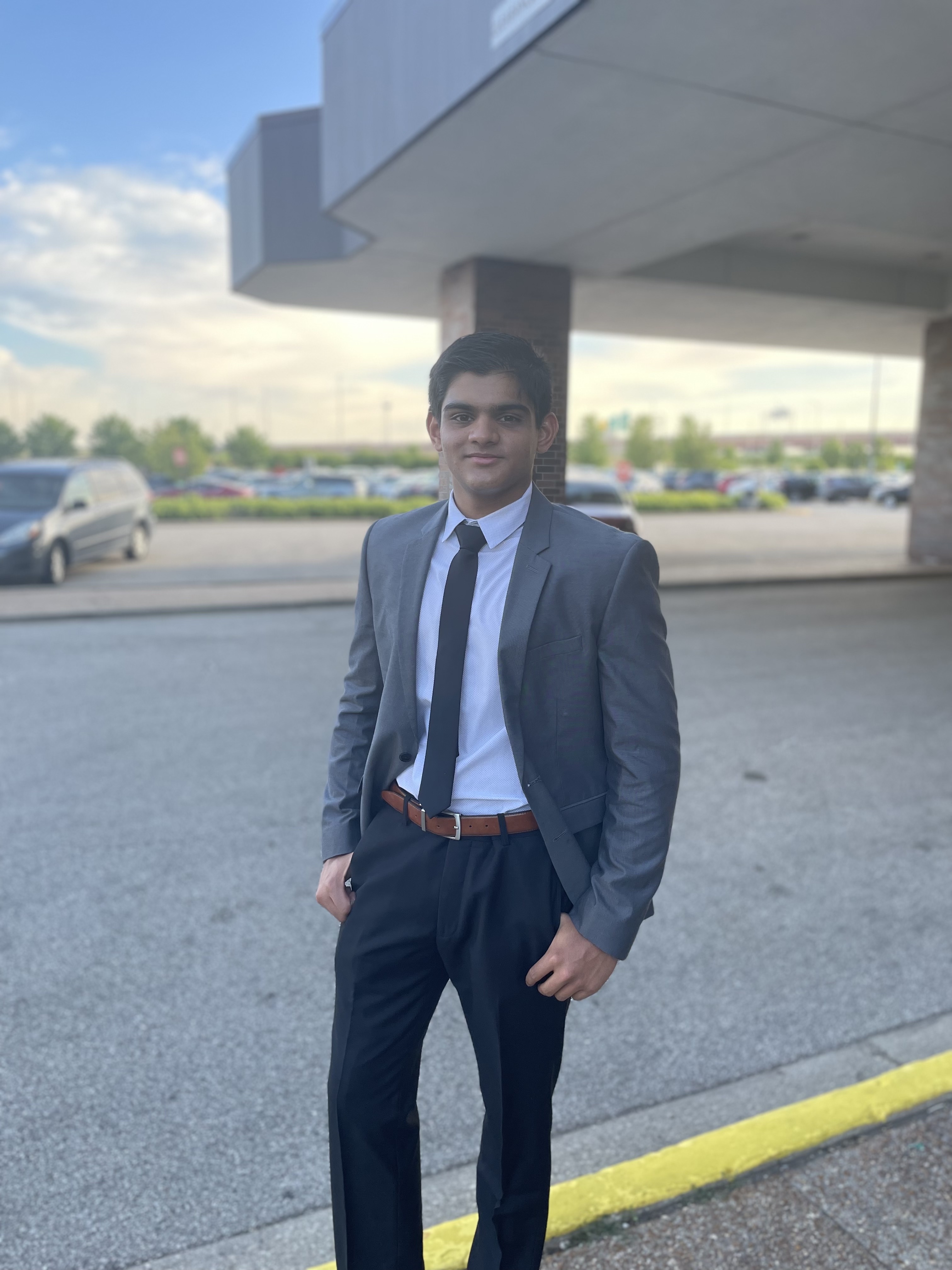 Person in a formal gray suit with a tie stands smiling under a building overhang, with a parking lot and cars visible in the background.