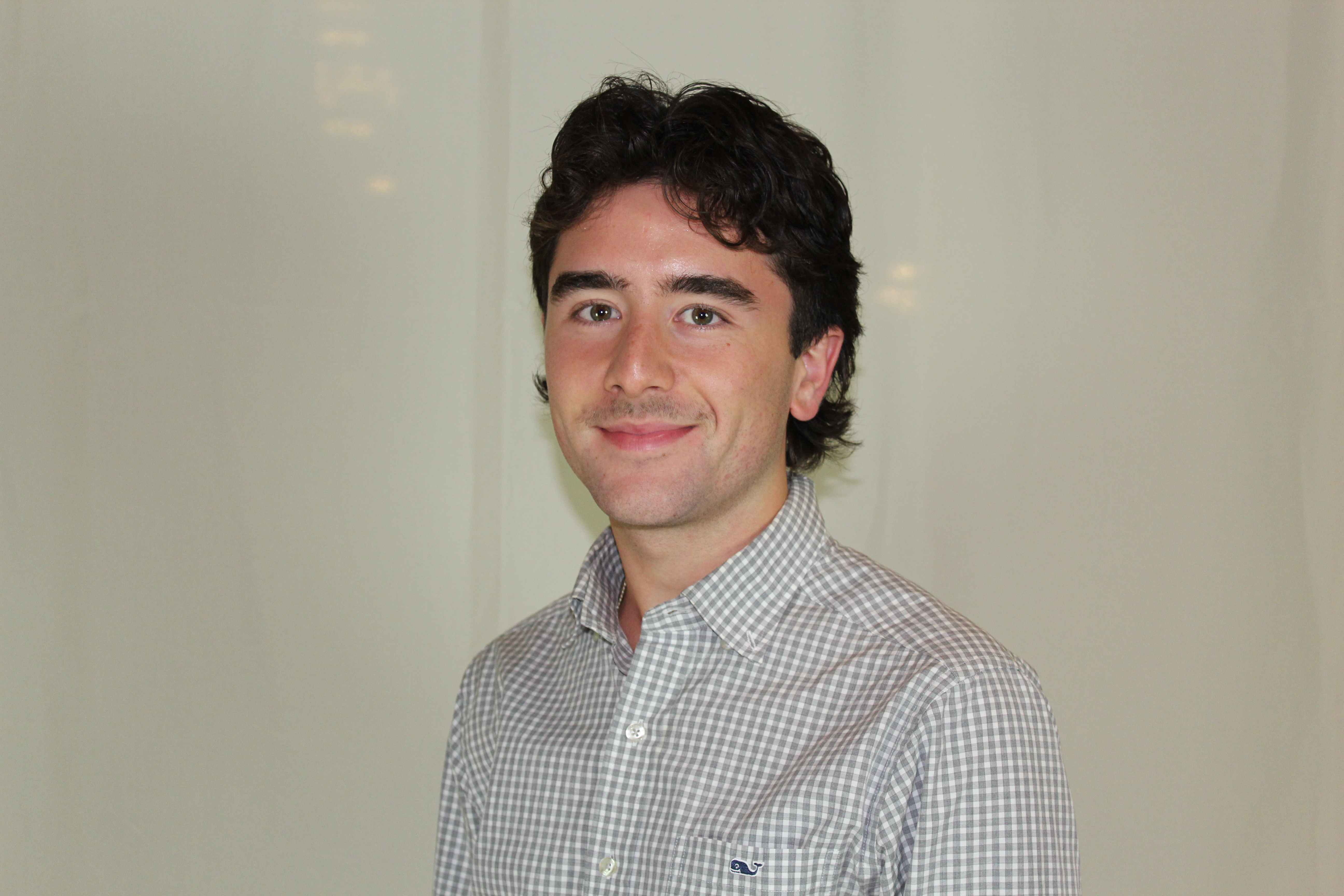 Portrait of a smiling person wearing a checkered shirt, standing indoors with a plain background.