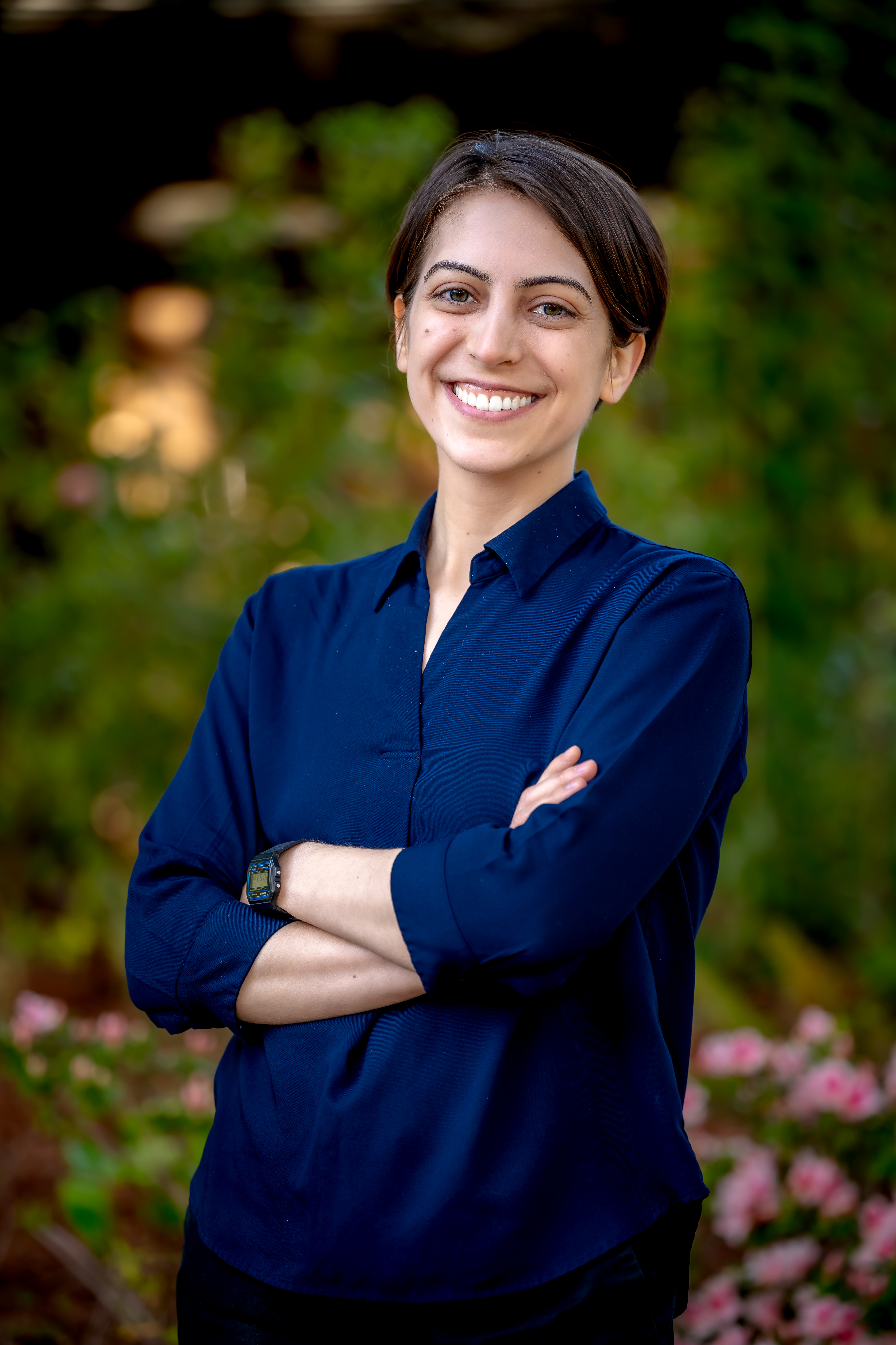 Person in a navy blue shirt smiling at the camera with arms crossed, standing in front of blurred greenery and flowers.