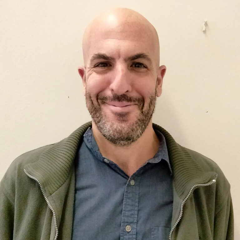 Smiling person with a shaved head, wearing a green shirt and olive jacket, standing against a plain wall.