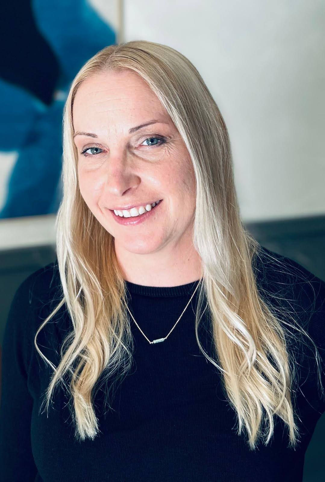 Person with long blonde hair smiling in front of a blue abstract art piece.