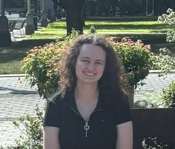 Person smiling, seated outdoors with green plants and trees in the background.