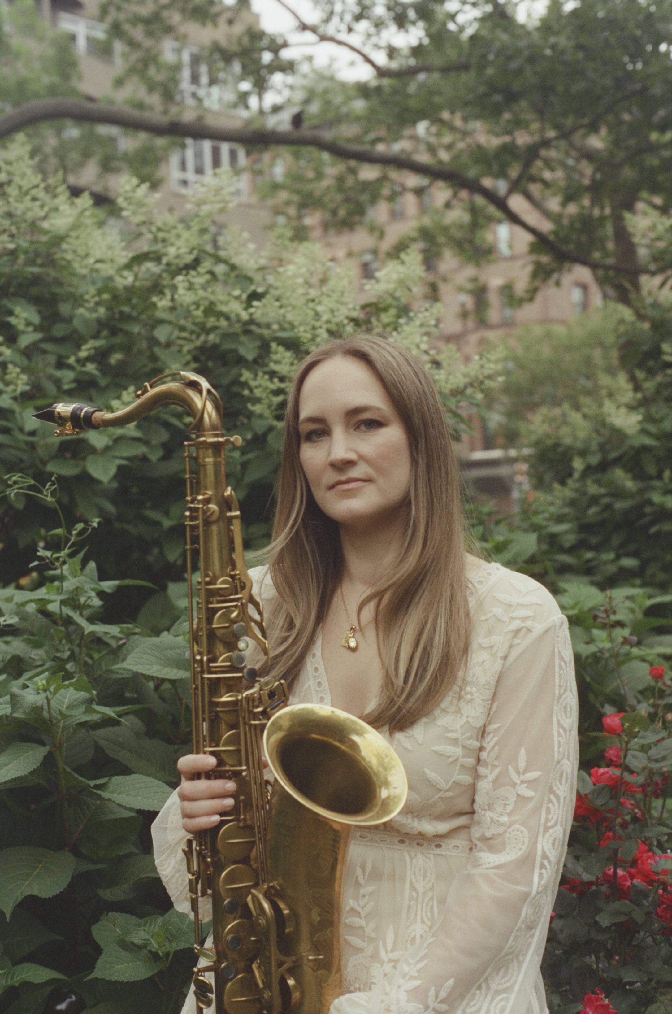 Individual holding a saxophone stands amidst lush greenery with red flowers and a brick building visible in the background.
