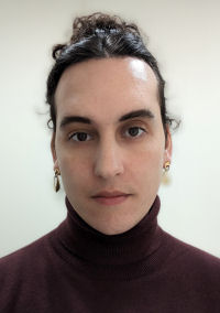 Portrait of a person with shoulder-length curly hair partially styled up, wearing a burgundy turtleneck and hoop earrings, against a plain light background.