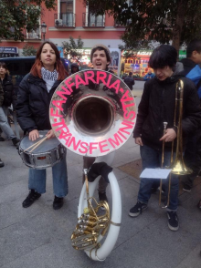 Transfeminist Music Band Three musicians playing in a street band, with a drum, a large horn, and a trombone. A banner on the horn reads "Fanfarria Transfeminista.
