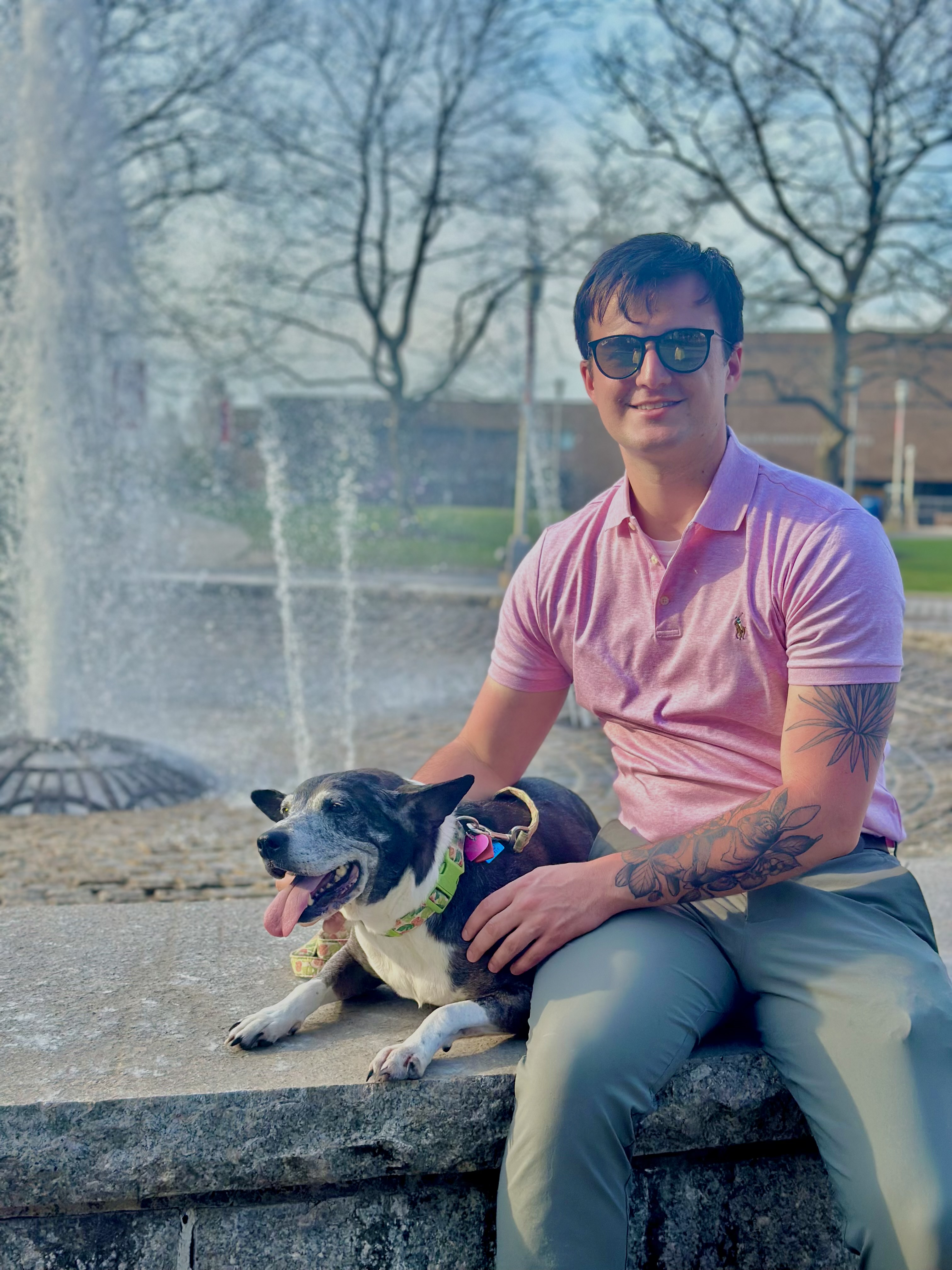 a smiling man wearing sunglasses sits on the edge of a fountain, petting a dog 