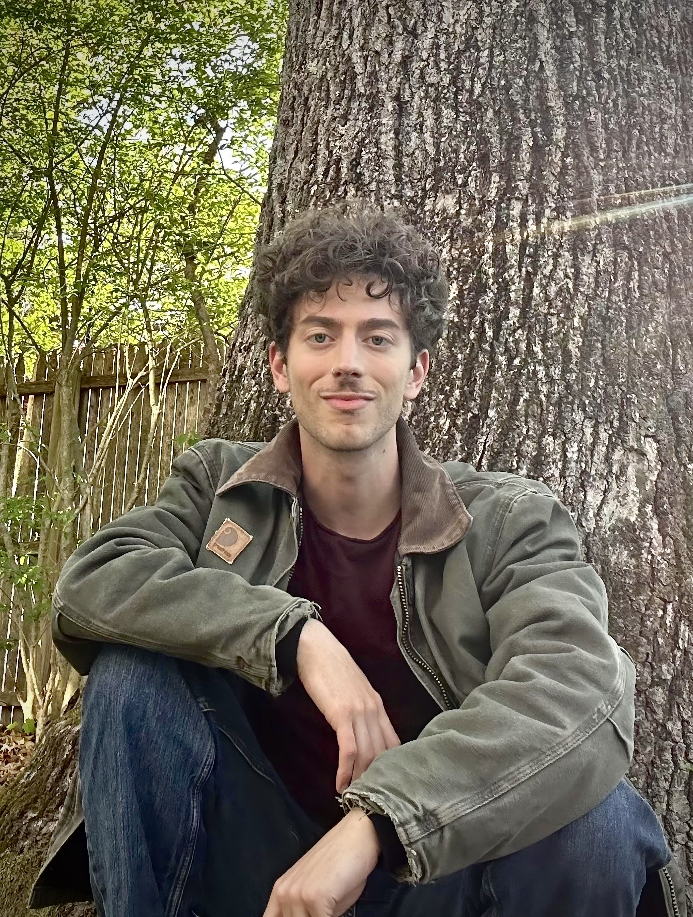 a smiling man with short hair sits in front of a tall tree 