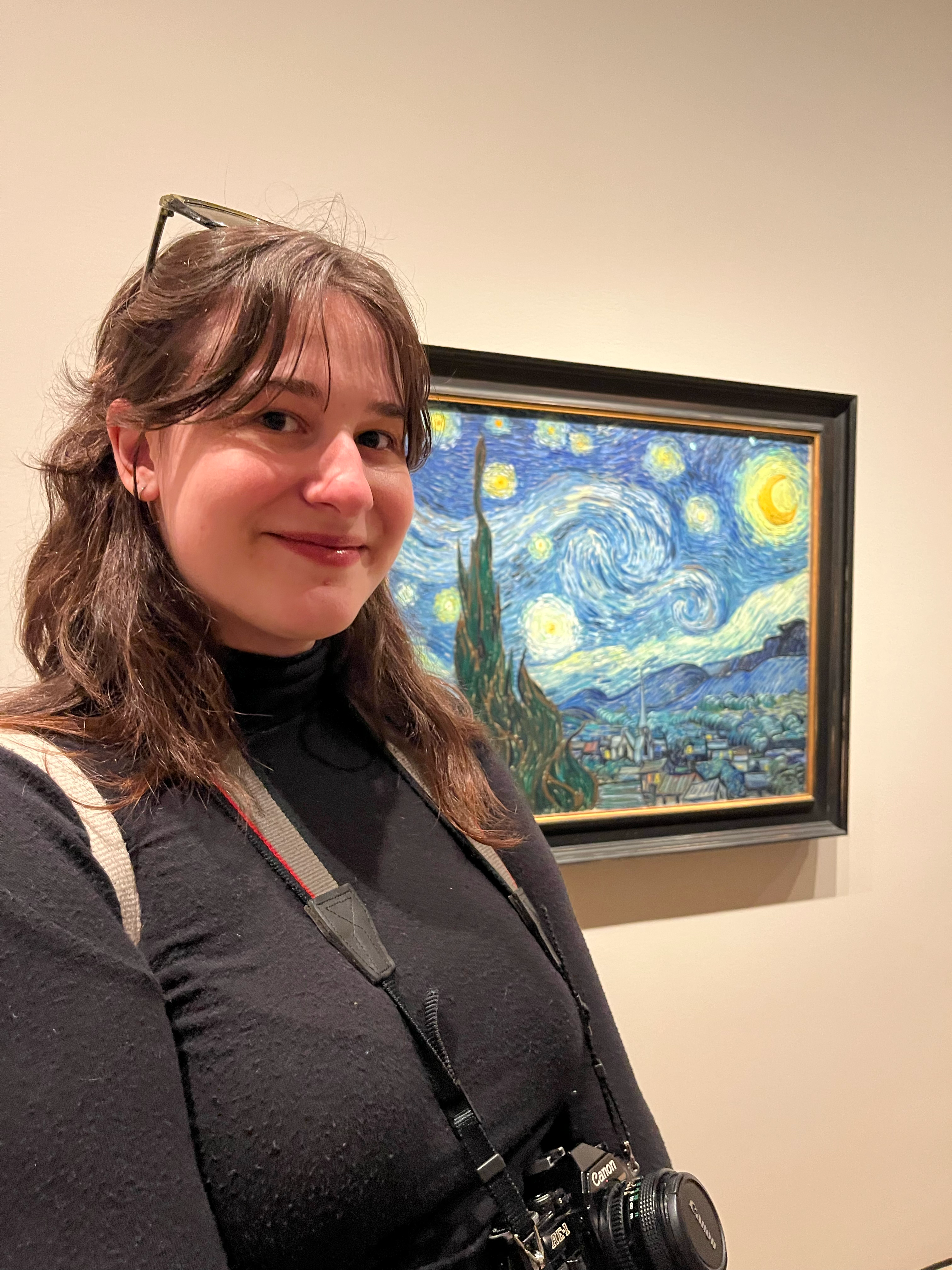 a smiling woman with sunglasses on her head and a camera around her neck stands in front of Starry Night in an art gallery