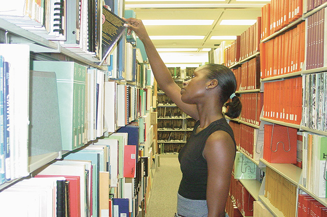 A person reaching for a book on a tall library shelf, surrounded by rows of colorful books. The scene conveys focus and curiosity in a well-lit space.
