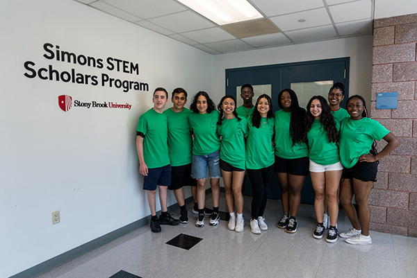 A diverse group of ten smiling young adults wearing green shirts stands together in a hallway under a sign reading "Simons STEM Scholars Program" at Stony Brook University.