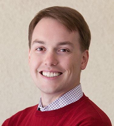 A professional headshot of a smiling man with short brown hair, wearing a red crewneck sweater over a white and purple checkered button-down shirt. He is posed against a neutral, textured off-white background.
