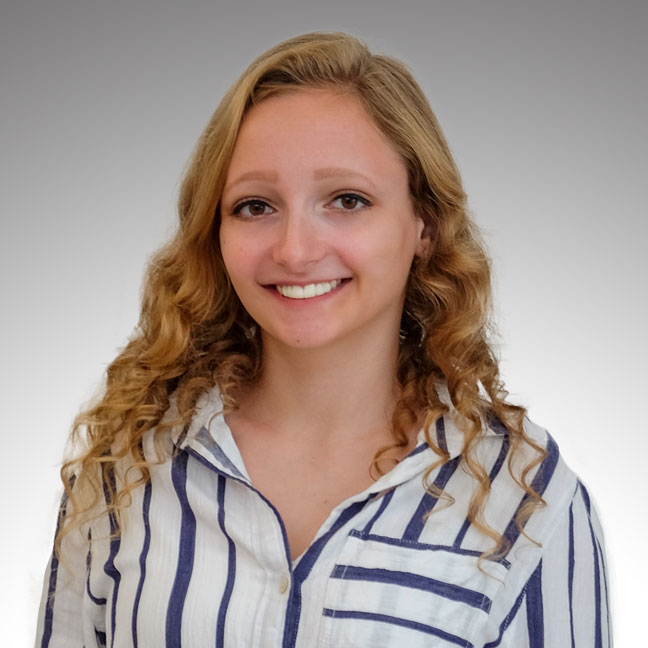 A professional headshot of a smiling woman with long, wavy blonde hair. She is wearing a white and navy blue vertically striped button-down shirt. The background is a smooth, neutral gradient of light grey to white.