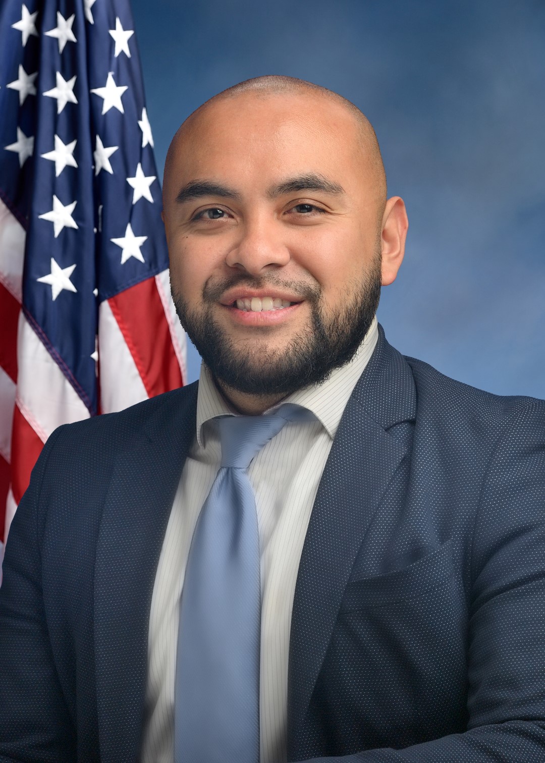 Steven Raga An individual dressed formally in a blue suit, white shirt, and light blue tie stands in front of a backdrop featuring the American flag and a gradient blue background.